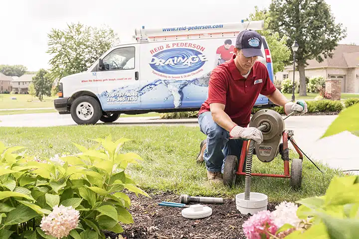 Reid & Pederson technician using a Ridgid K-1500 sewer machine to perform outdoor drain cleaning in Crete, IL. A branded company van is parked in the background, highlighting their professional sewer and drain services.